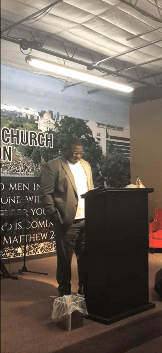 Person speaking at a podium with a church backdrop and Bible verse visible behind.