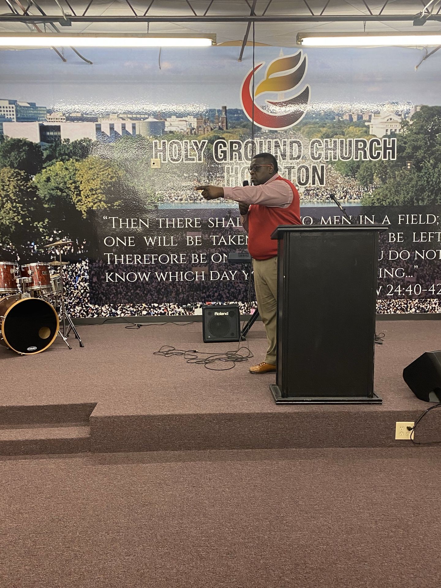 Speaker at podium in church with mural and drum set in background, pointing and speaking.