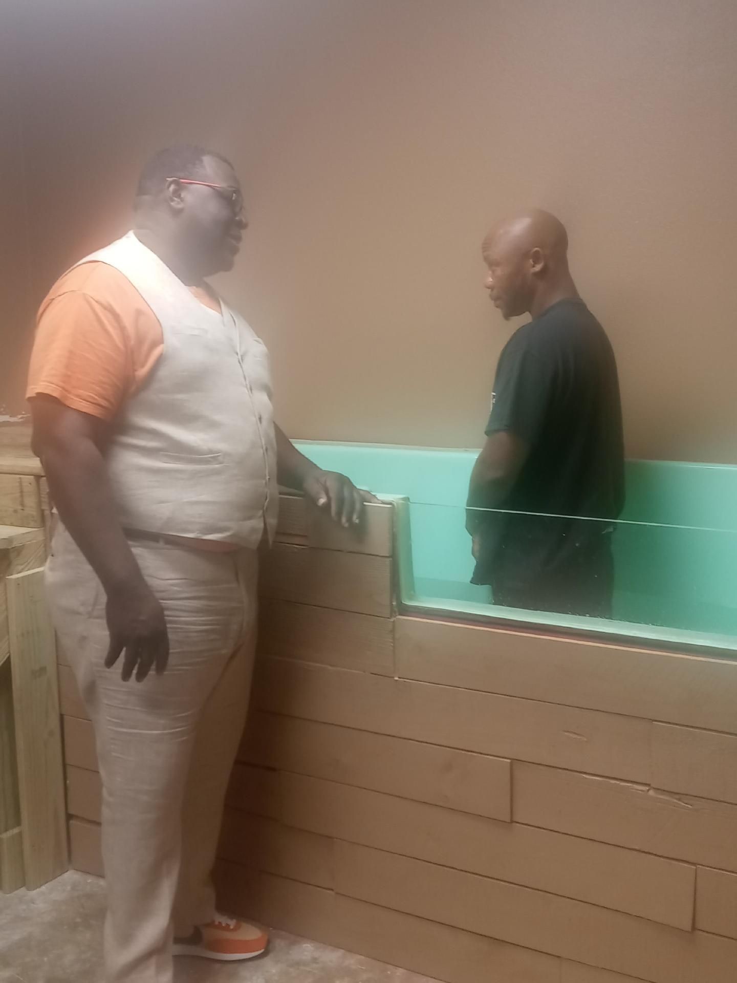Two men having a conversation near a teal partition in a room with brown walls.